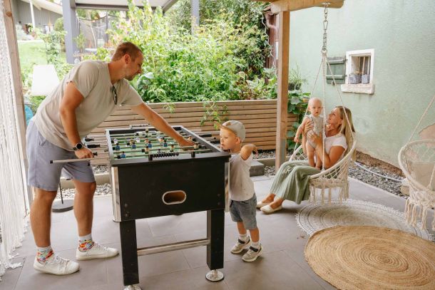 The father plays with his son at the foosball table while the mother and baby sit in the swing