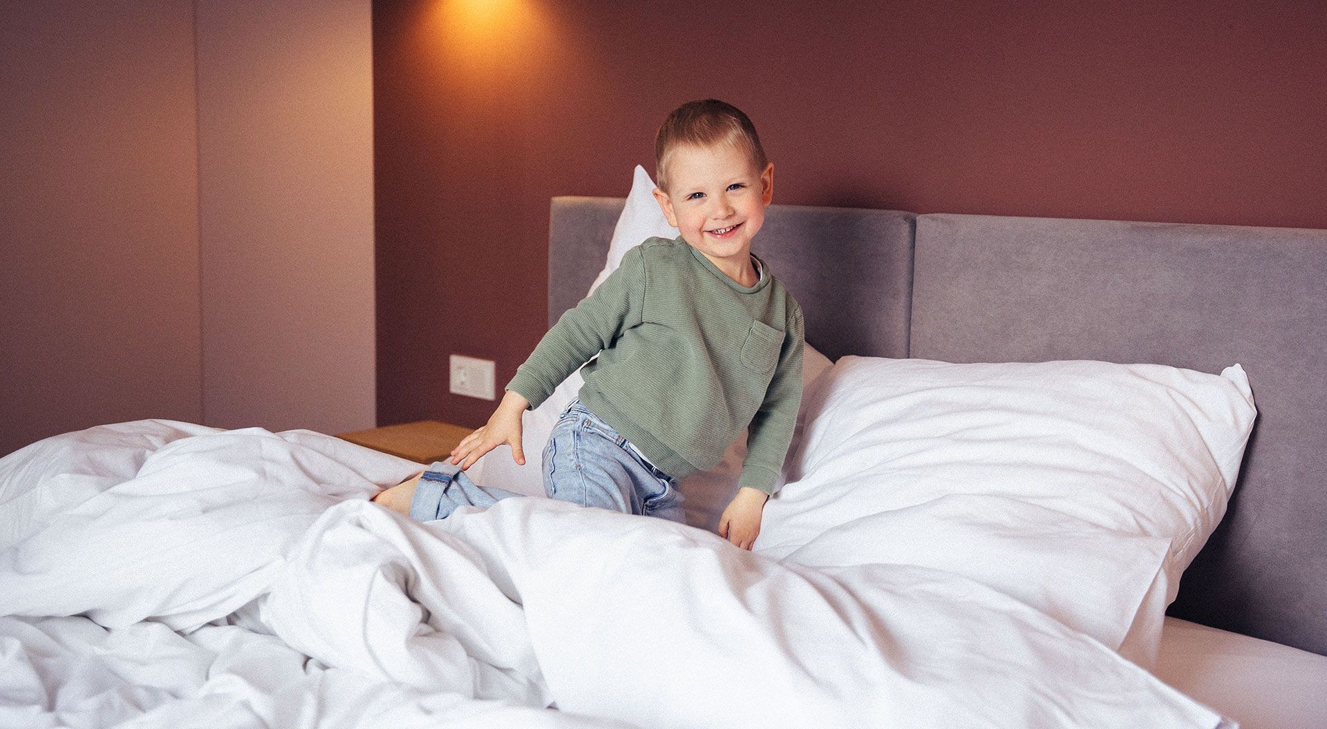 A little boy plays on the bed and grins at the camera